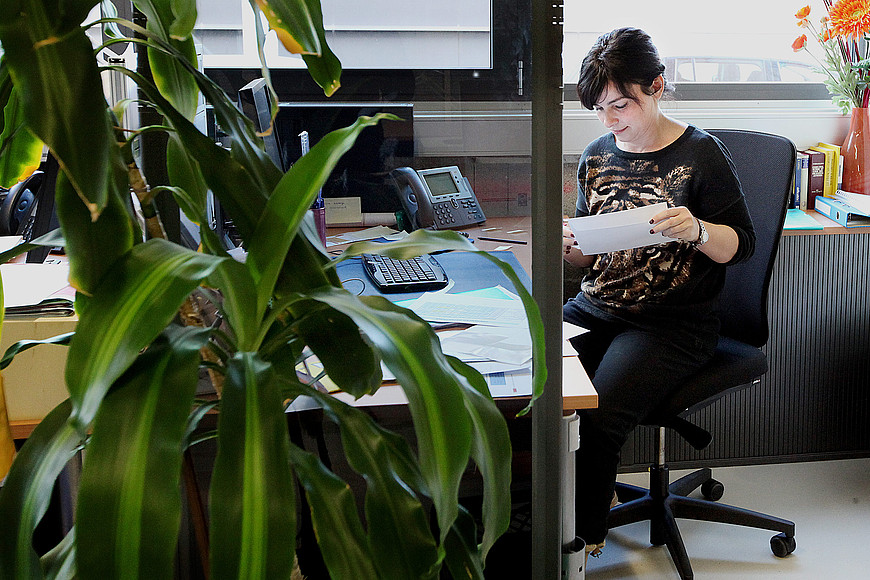 Une femme est assise à son bureau, elle est en train de fermer une enveloppe, une grande plante verte est dans le coin à gauche de l'image